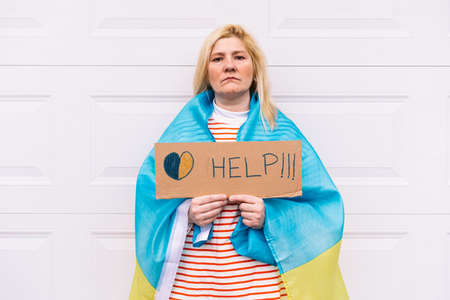 Ukrainian woman with blonde hair and a serious gesture, with a blue and yellow Ukrainian flag holding a sign with a yellow and blue heart that reads: 'Help', on the street. Concept Ukraine war, invasion, protest, patriotism.の写真素材