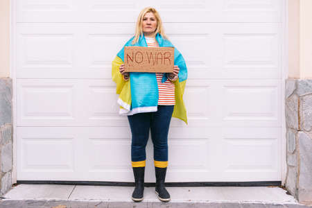 Ukrainian woman with blonde hair and a serious gesture, with a blue and yellow Ukrainian flag with a sign that reads: 'No War', on the street. Concept Ukraine war, invasion, protest, patriotism.の写真素材
