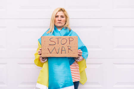 Ukrainian woman with blonde hair and a serious gesture, with a blue and yellow Ukrainian flag with a sign that reads: 'Stop War', on the street. Concept Ukraine war, invasion, protest, patriotism.の写真素材