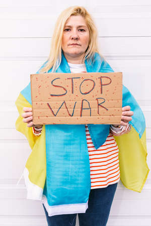 Ukrainian woman with blonde hair and a serious gesture, with a blue and yellow Ukrainian flag with a sign that reads: 'Stop War', on the street. Concept Ukraine war, invasion, protest, patriotism.の写真素材