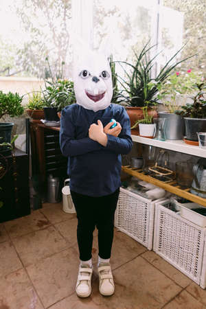 Girl celebrating Easter with a rabbit mask, arms crossed and some chocolate eggs in a glassed-in area surrounded by plants. Easter celebration and costume concept.の写真素材