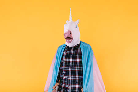 Person dressed in a unicorn mask with a checkered shirt holding the flag of the transsexual collective, on a yellow background. Concept of gay pride, transsexuality, and lgtbq rights.の写真素材