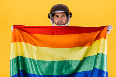 Gay man dressed as an astronaut with a helmet and silver suit with a rainbow lgtbi collective flag, on a yellow background. Gay, homosexual, trans, rights and gender pride concept.の写真素材