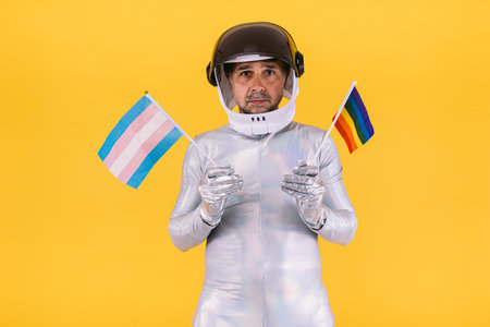 Gay man dressed as an astronaut with a helmet and silver suit, with flags of the lgtbi and trans collective, on a yellow background. Gay, homosexual, trans, rights and gender pride concept.の写真素材
