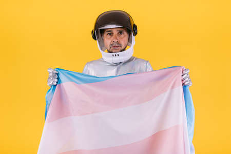 Gay man dressed as an astronaut with a helmet and silver suit, with the flag of the trans collective, on a yellow background. Gay, homosexual, trans, rights and gender pride concept.の写真素材