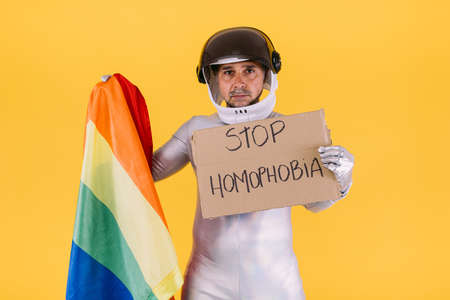 Gay man dressed as an astronaut with a helmet and silver suit, with the flag of the lgtbi collective holding a sign that reads: 'stop homophobia', on a yellow background.の写真素材