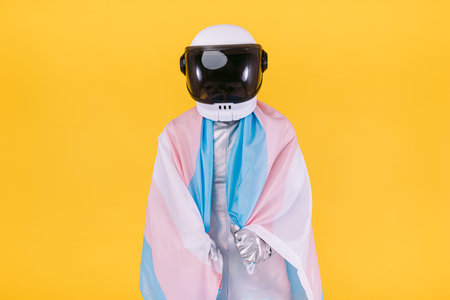Gay man dressed as an astronaut with a helmet and silver suit, holding a flag of the trans...