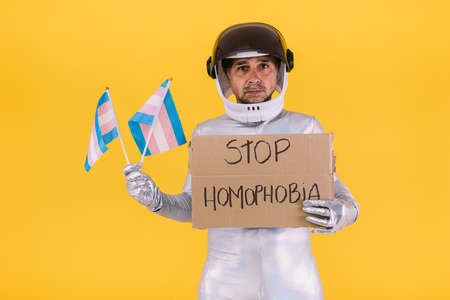 Gay man dressed as an astronaut with a helmet and silver suit, holding two small flags of the trans collective and a sign that reads: Stop homophobia, on a yellow background.の写真素材