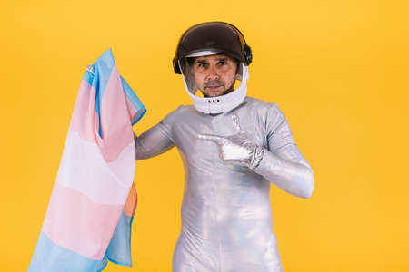 Gay man dressed as an astronaut with a helmet and silver suit, pointing to a flag of the trans collective, on a yellow background. Gay, homosexual, trans, rights and gender pride concept.の写真素材