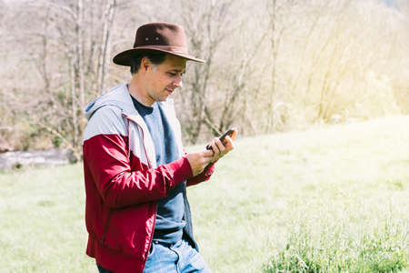 Entrepreneurial self-employed man wearing a hat, looking at his mobile phone while relaxing in the field, on a meadow. Concept of work, enjoy, relax, Internet, technology and connectivity.の写真素材