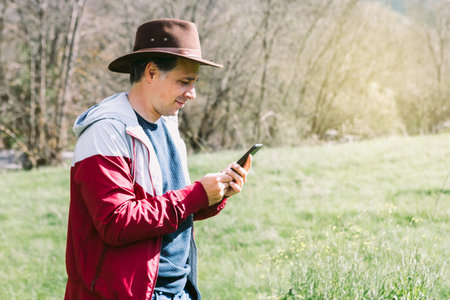 Entrepreneurial self-employed man wearing a hat, looking at his mobile phone while relaxing in the field, on a meadow. Concept of work, enjoy, relax, Internet, technology and connectivity.の写真素材
