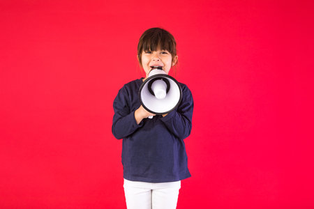 Black-haired girl in a blue sweater and white pants, speaking through a white megaphone, on a red background.の写真素材