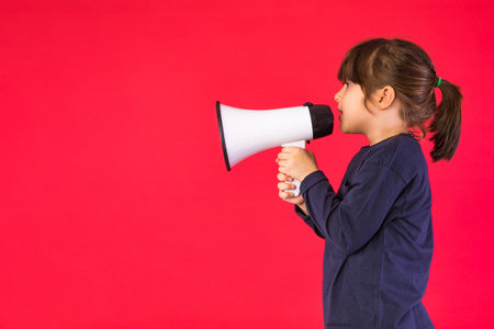 Black-haired girl in a blue sweater and white pants, shouting through a white megaphone, on a red background.の写真素材