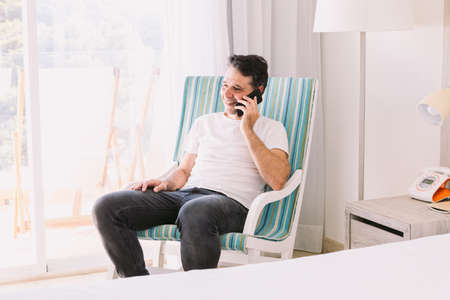 Young man sitting in a rocking chair in his bedroom, talking on the cell phone and smiling, with the light coming through the window. Concept of working, vacation, connection and smartphone.の写真素材