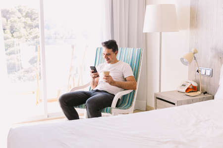 Young man sitting in a rocking chair in his bedroom, looking at the mobile and holding a drink, with the light coming through the window. Concept of working, vacation, break, breakfast, cafe.の写真素材