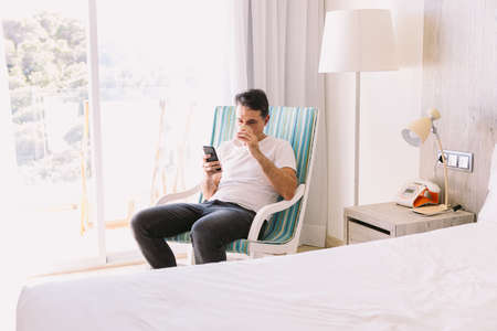 Young man sitting in a rocking chair in his bedroom, looking at his cell phone and drinking a coffee, with the light coming through the window. Concept of working, vacation, break, breakfast, cafe.の写真素材