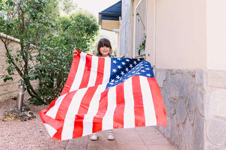 Black-haired girl holding and waving a United States flag, in the garden of her house. Concept of celebration, independence day, United States of America, 4th of July, patriotism and American pride.の写真素材