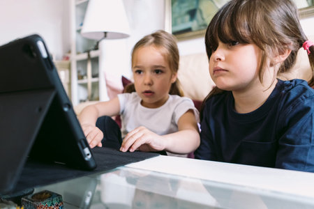 Two little girls attentively watching a video on a tablet in their living room. Concept of...