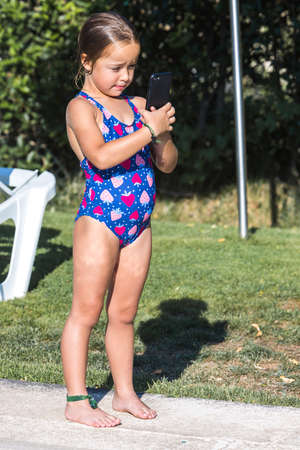Little girl in a swimsuit, sitting on the edge of the pool, looking at the mobile phone. Childhood, children, smartphone, technology, vacation, internet and fun concept.の写真素材