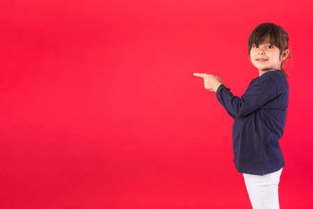 Smiling brunette haired girl, wearing blue sweatshirt and white pants, pointing fingers to the side with copy space, on red background. Concept of call center, call, speak and listen.の写真素材
