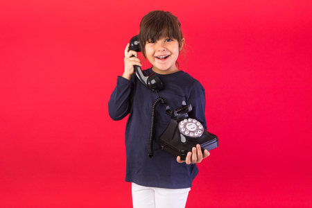 Smiling brunette-haired girl, wearing blue sweatshirt and white pants, holding retro phone and talking on the earpiece on red background. Concept of call center, call, speak and listen.の写真素材