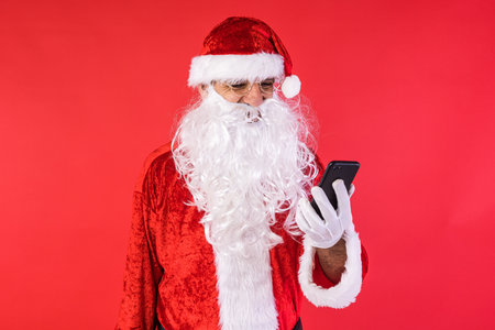 Man dressed as Santa Claus, consulting his mobile phone, on red background. Christmas, celebration, gifts, consumerism and happiness concept.の写真素材