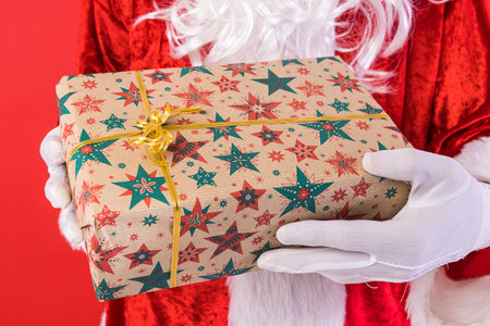 Detail of the hands of a man dressed as Santa Claus with gloves holding a gift, on a red background. Christmas, celebration, gifts, consumerism and happiness concept.の写真素材