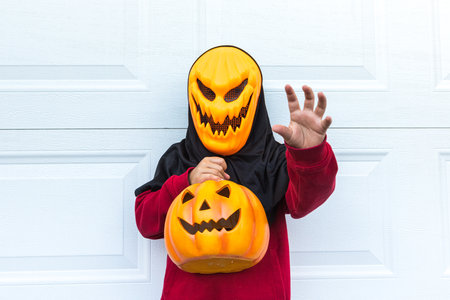 A child girl wearing a scary Halloween pumpkin mask, holding a pumpkin, scaring with her hand, over a white garage door. Concept of celebration, costume, carnival, terror, fear and autumn.の写真素材