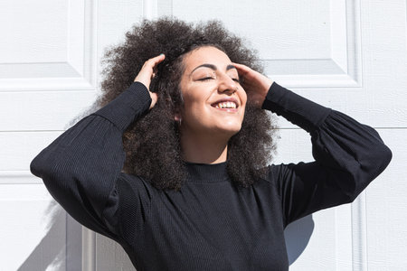 Young white woman with afro-style hair, dressed in black, leaning against a white door, smiling,...