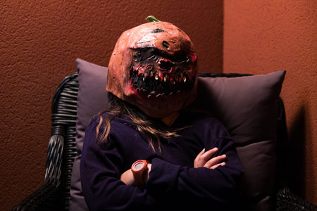 A young person wearing a Halloween pumpkin mask, arms crossed, sitting in a chair on a terrace.