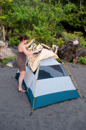 Woman demonstrates how to pitch a 2 person tent with a rain fly on a black sand beach campsite.の写真素材