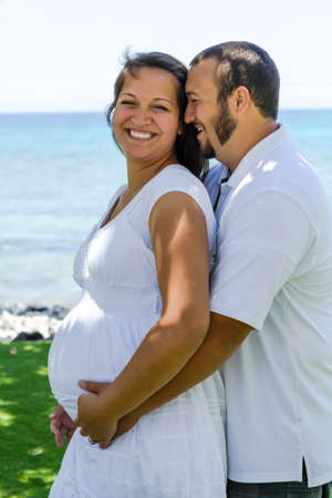 Husband whispering to his pregnant wife while holding her close next to the ocean の写真素材