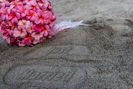 Just Married stamped in the sand next to a bouquet of pink plumeria flowers の写真素材