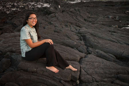 Barefoot young lady happily sitting on the seaside lavarock at sunset in Hawaii の写真素材