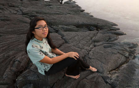 Barefoot young lady wearing glasses, sitting on lava rock at the waterの写真素材