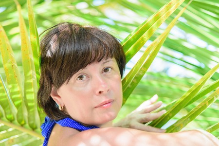brunette middle-aged woman in a bathing suit among the leaves of palm treesの写真素材