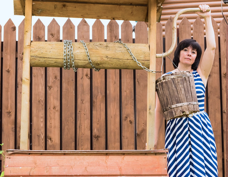 middle-aged brunette woman standing at the village well with a wooden bucketの写真素材
