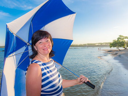 Brunette middle-aged woman on the beach in a beautiful dress with striped umbrellaの写真素材