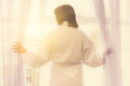 brunette middle-aged woman at the window at dawn wins sunlightの写真素材