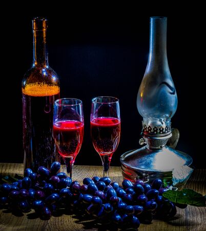 a bottle of red wine and two glasses against a background of black grapes in the dim light from a kerosene lampの写真素材