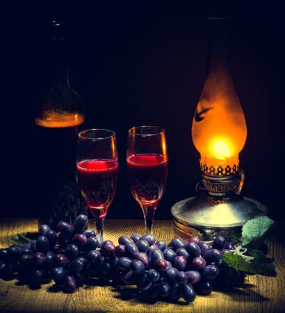 a bottle of red wine and two glasses against a background of black grapes in the dim light from a kerosene lampの写真素材