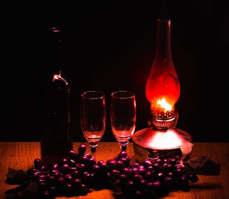 a bottle of red wine and two glasses against a background of black grapes in the dim light from a kerosene lampの写真素材