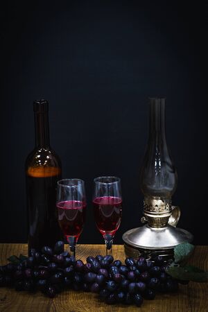 a bottle of red wine and two glasses against a background of black grapes in the dim light from a kerosene lampの写真素材