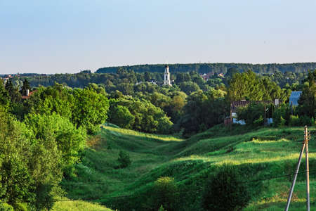 Rural landscape ravine overgrown with green grass and trees. Bell tower of Christian church in the backgroundの写真素材