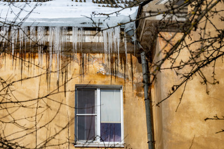 Big beautiful icicles hang from the snow-covered roof in front of the windows of the house. Snow on...
