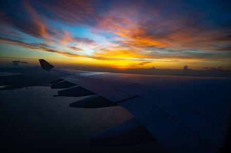 A plane wing is flying over the ocean with a beautiful sunset in the background. The sky is filled with clouds and the sun is setting, creating a serene and peaceful atmosphereの写真素材