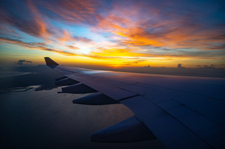 A plane wing is flying over the ocean with a beautiful sunset in the background. The sky is filled with clouds and the sun is setting, creating a serene and peaceful atmosphereの写真素材