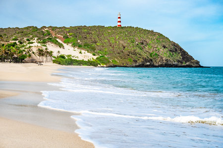 A beach with a lighthouse in the distance. The beach is empty and the water is calm. The sky is clear and the sun is shiningの写真素材