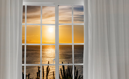 Window with a curtain that has a picture of a sunset on it. The sun is setting and the water is calmの写真素材
