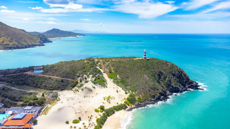 Beach with a lighthouse on a hill. The water is blue and the sky is clear. There are some buildings in the background filmed from a droneの写真素材